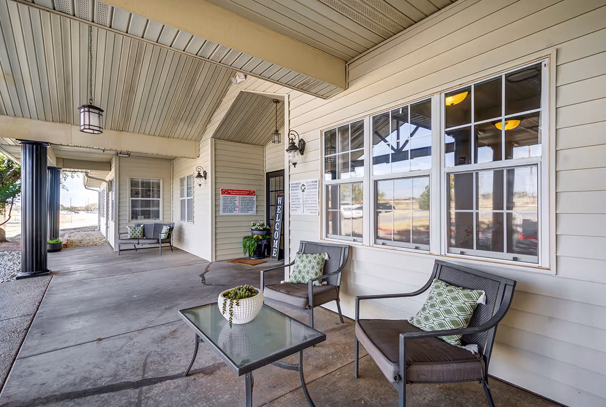 Covered outdoor seating area at the entrance of a facility with two cushioned chairs and a glass-top table with a potted plant. The building has beige siding, large windows, hanging lanterns, and a welcome sign near the door.