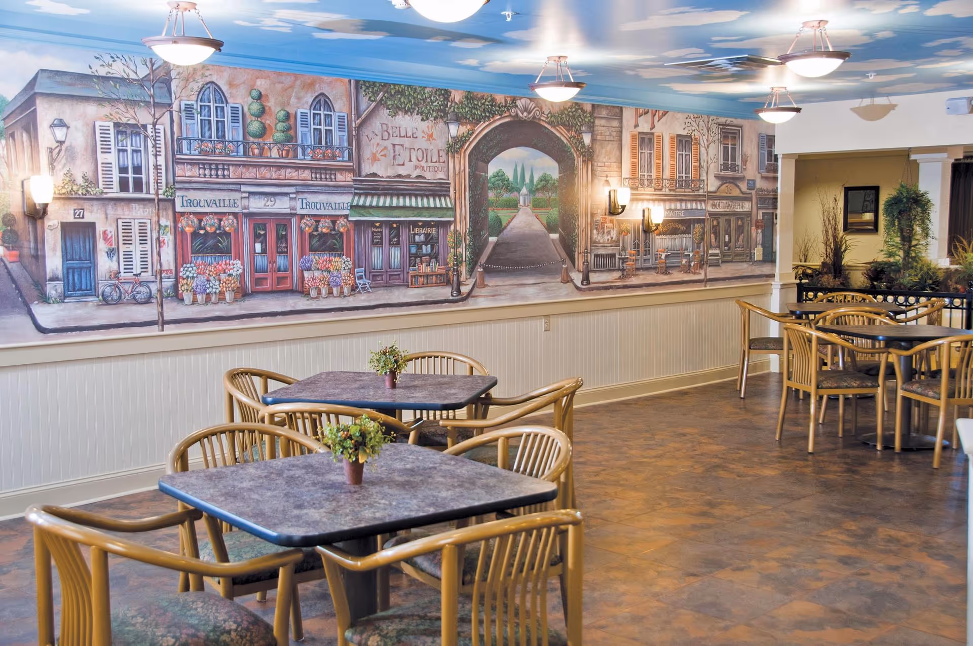 Interior dining area with several tables and chairs. Each table has a small potted plant. The wall features a large mural depicting a quaint European street scene with shops and an archway leading to a garden path. Ceiling lights illuminate the space.
