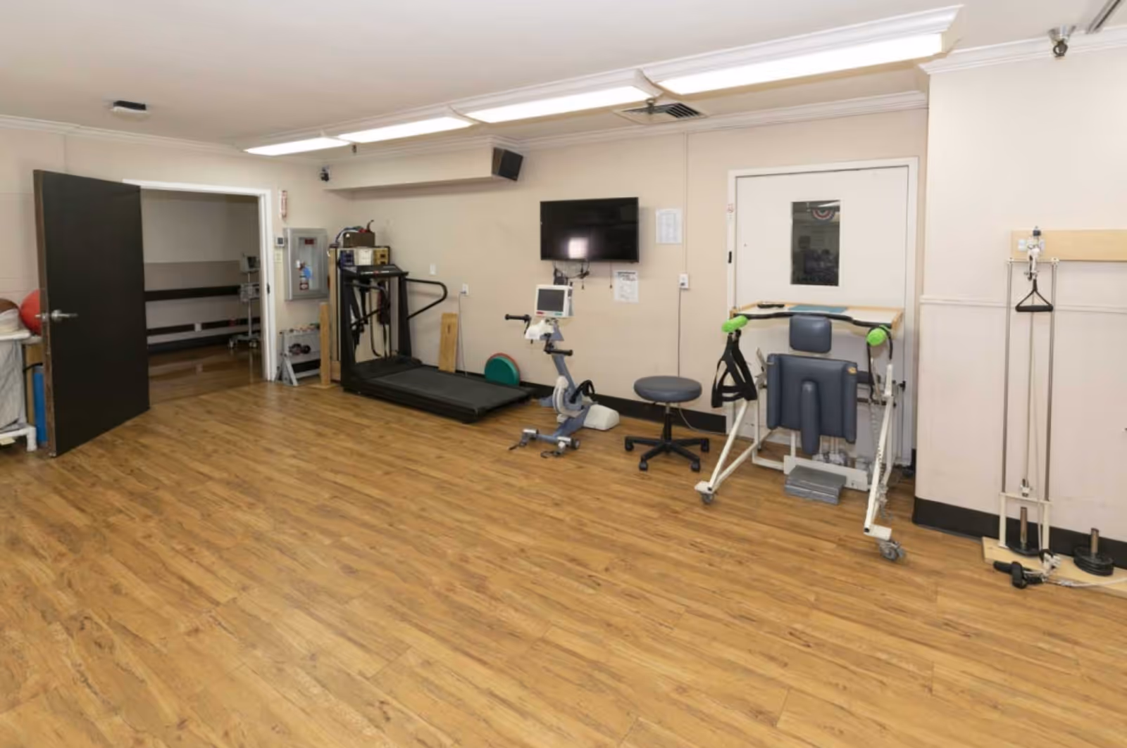 A rehabilitation room with wooden flooring, a treadmill, an exercise bike, a stool, a TV mounted on the wall, and various physical therapy equipment. There is an open door leading to another room with benches.