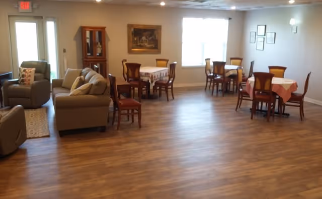 A spacious room with wooden flooring featuring several small dining tables covered with tablecloths and surrounded by wooden chairs. There are also two armchairs and a sofa arranged near the left side of the room. The walls are light-colored with framed pictures and a window letting in natural light.