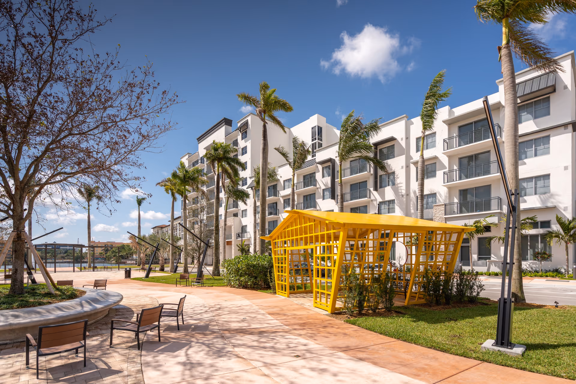 Outdoor area of Overture Doral featuring a modern white multi-story building with balconies, tall palm trees, a curved paved walkway with benches, and a bright yellow open pavilion structure under a blue sky with a few clouds.