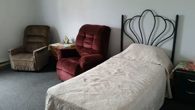 A simple bedroom with a single bed covered in a light-colored bedspread, a black metal headboard, a red cushioned armchair, a brown cushioned armchair, and two small side tables with decorative items on them. The room has plain white walls and a carpeted floor.