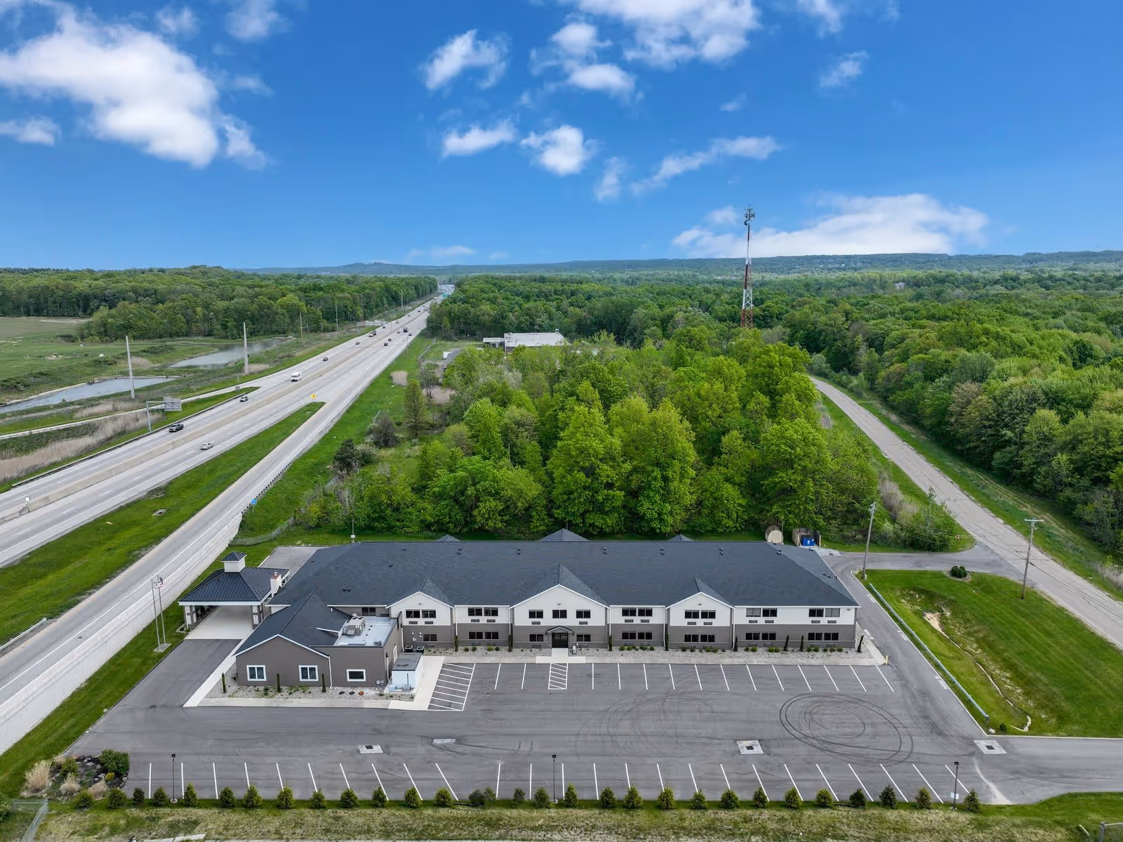 Aerial view of a single-story senior living facility building with a large empty parking lot in front, surrounded by green trees and grass. A highway runs parallel to the left side of the building, and a tall communication tower is visible in the background under a partly cloudy blue sky.