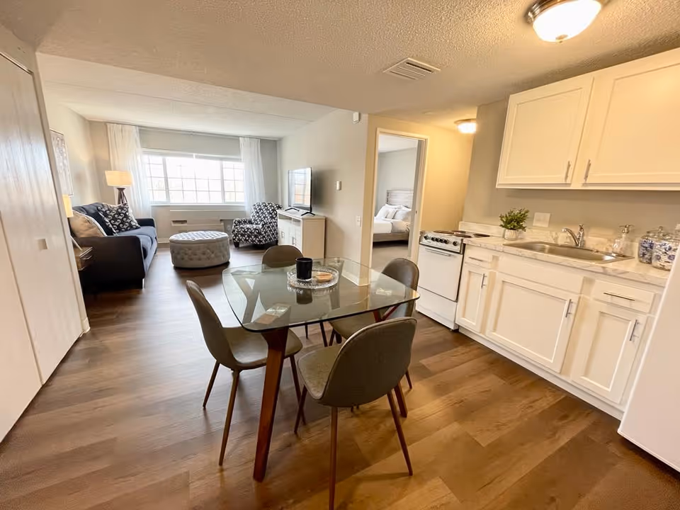 A bright and cozy living space featuring a small kitchen area with white cabinets, a sink, and a stove on the right. In the center, there is a glass dining table with four chairs. The living room area at the back has a dark sofa, a patterned armchair, a round ottoman, and a large window with white curtains letting in natural light. A doorway leads to a bedroom with a bed visible.