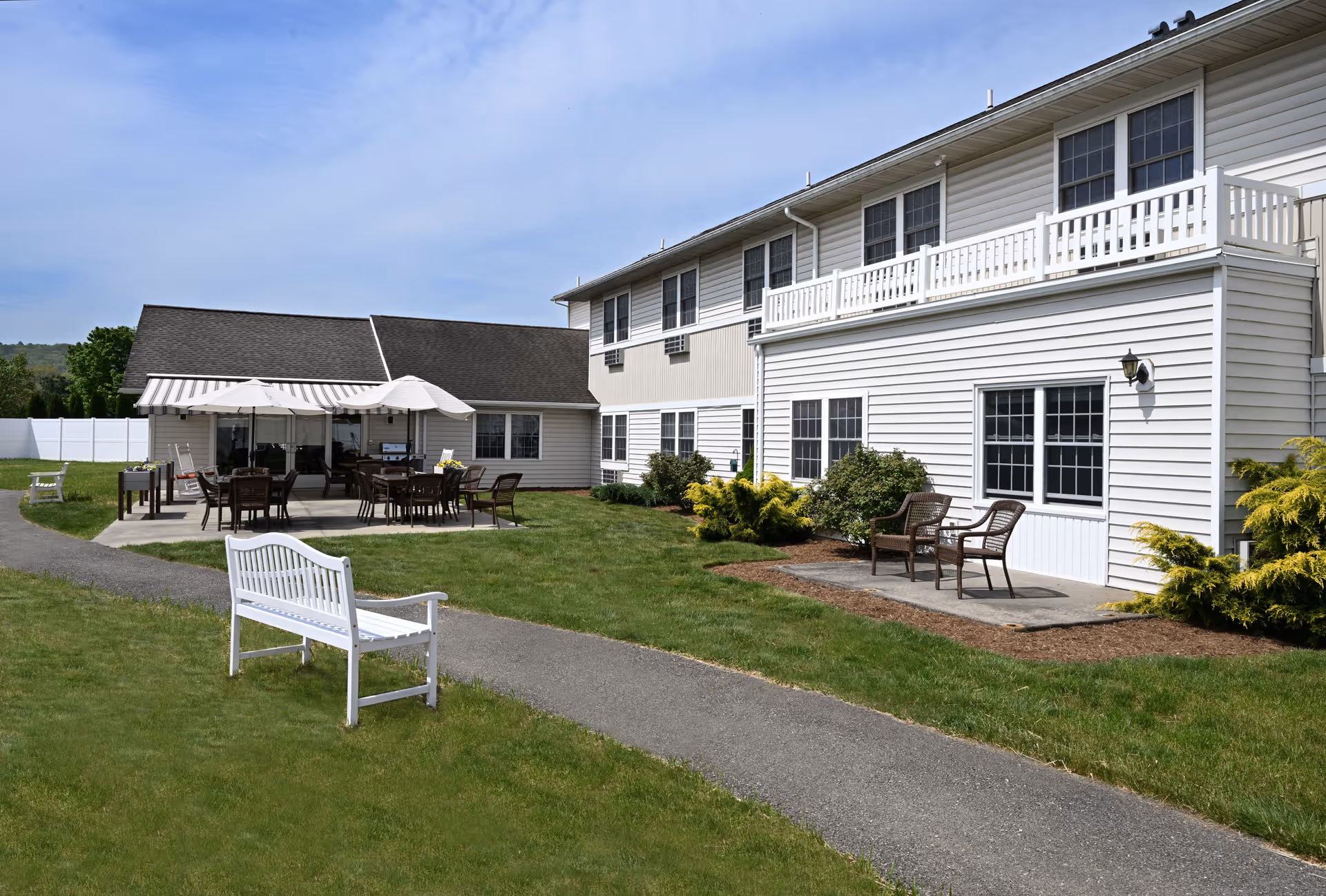 Outdoor patio area at Providence Place Senior Living of Pine Grove with white benches, tables with umbrellas, chairs, and a paved walkway surrounded by green grass and shrubs next to a two-story white building.