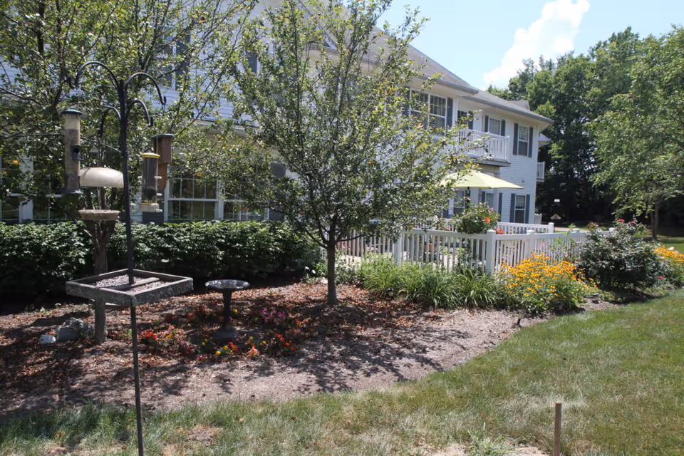 A garden area with a bird feeder and bird bath in front of a white two-story building with balconies and a patio with a yellow umbrella. The garden has various plants and flowers, and there are trees and bushes surrounding the area under a partly cloudy sky.