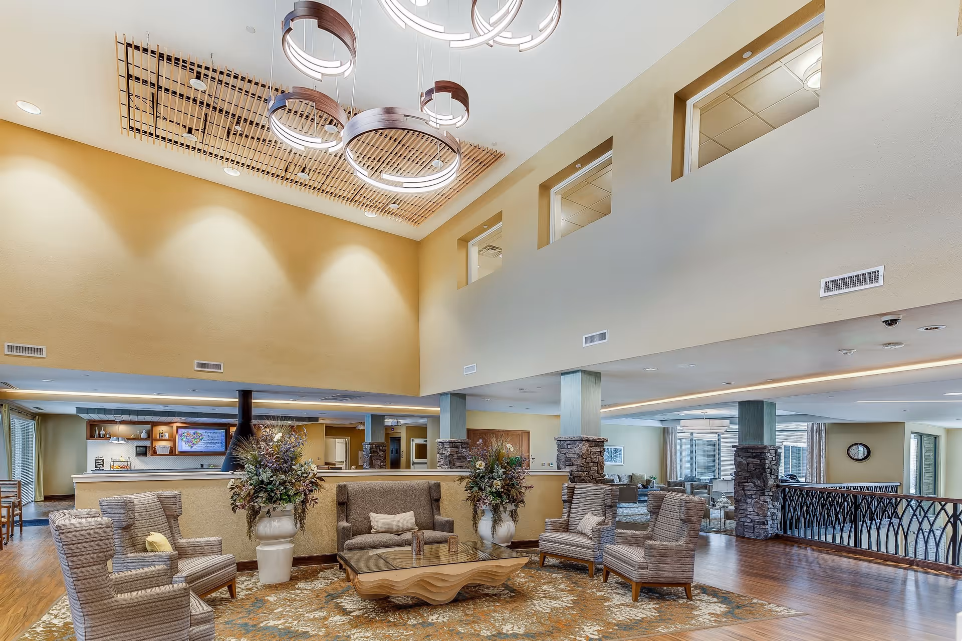 Spacious and well-lit common area in a senior living facility featuring a high ceiling with modern circular light fixtures, comfortable seating including armchairs and a sofa arranged around a wooden coffee table on a patterned rug, decorative large flower vases, and a railing overlooking a lower level.