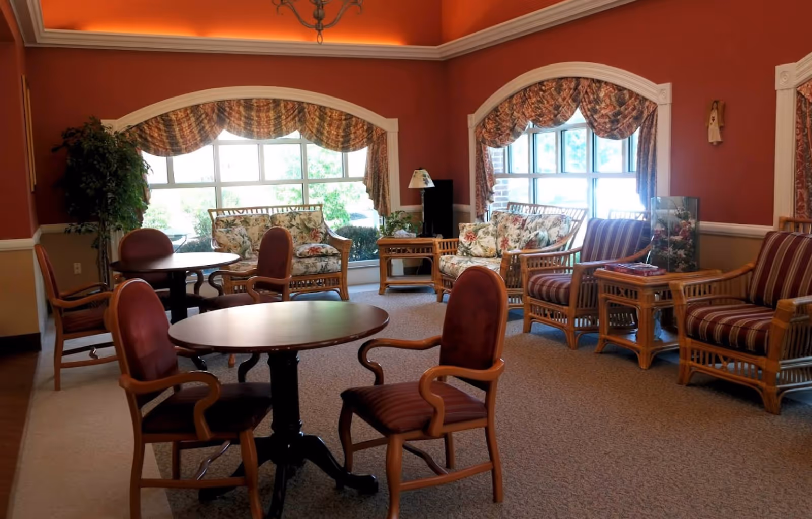 A cozy common room with round tables, upholstered chairs, wicker sofas and large arched windows with patterned valances.