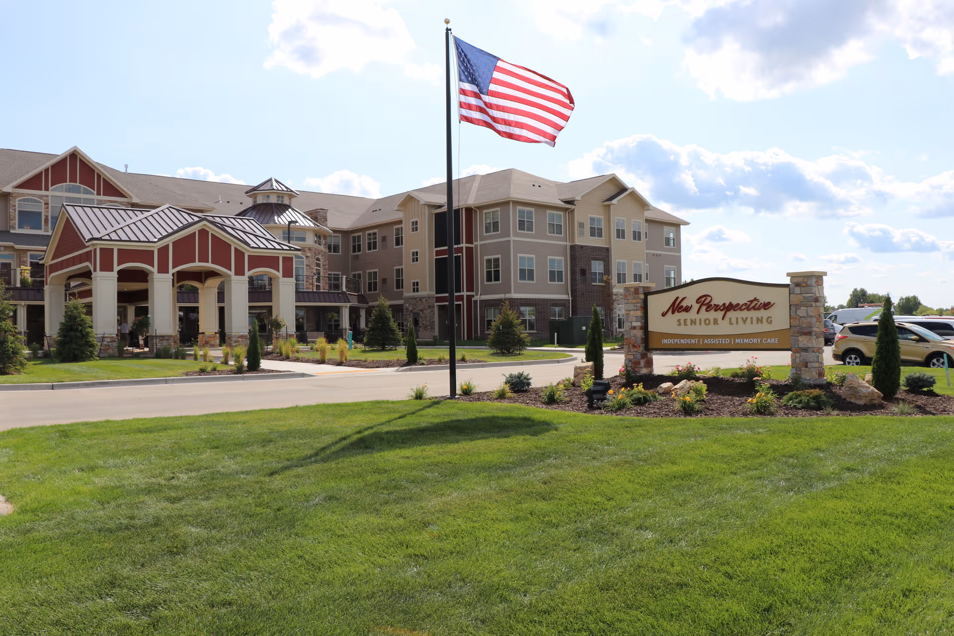 Front exterior of New Perspective Senior Living featuring the entrance, landscaping, an American flag, and the facility sign on a sunny day.