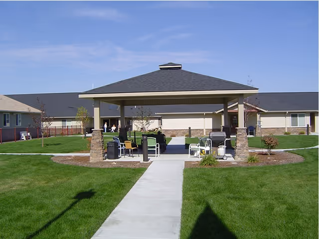 Outdoor covered pavilion with seating and grills in a grassy courtyard area surrounded by single-story buildings under a clear blue sky.