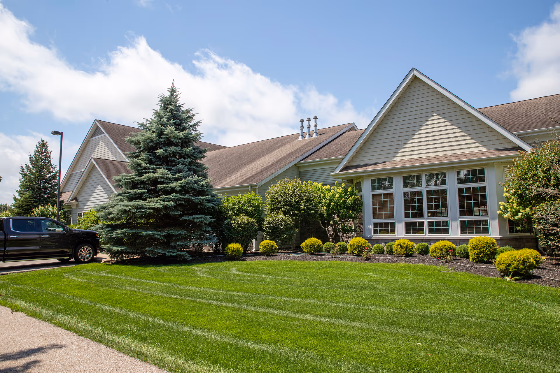 Exterior view of a single-story building with beige siding and a brown roof, surrounded by well-maintained green grass, bushes, and a large evergreen tree. A black pickup truck is parked on the driveway to the left. The sky is partly cloudy with blue patches.
