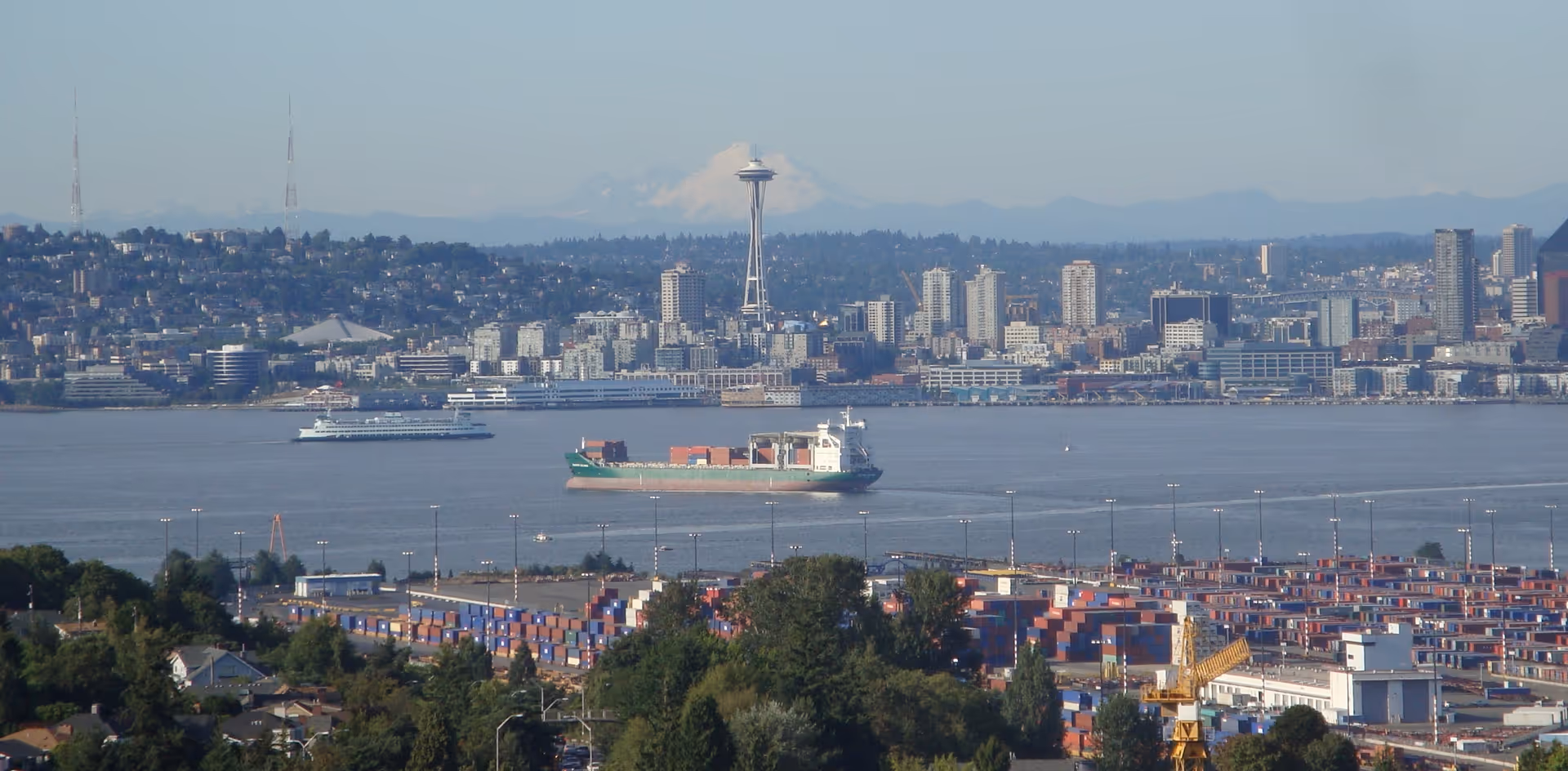 View of a city skyline with the Space Needle prominently visible, a large body of water with a cargo ship and a ferry, and a foreground of trees and shipping containers.