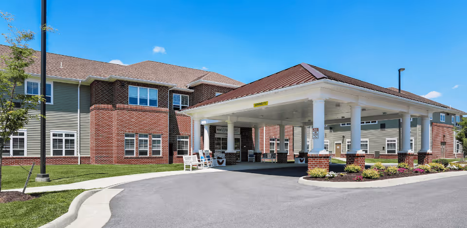 Exterior view of a senior living facility named Harmony at Martinsburg, showing a large covered entrance with white columns, brick and siding exterior walls, a paved driveway, and landscaped areas with grass and flowers under a clear blue sky.
