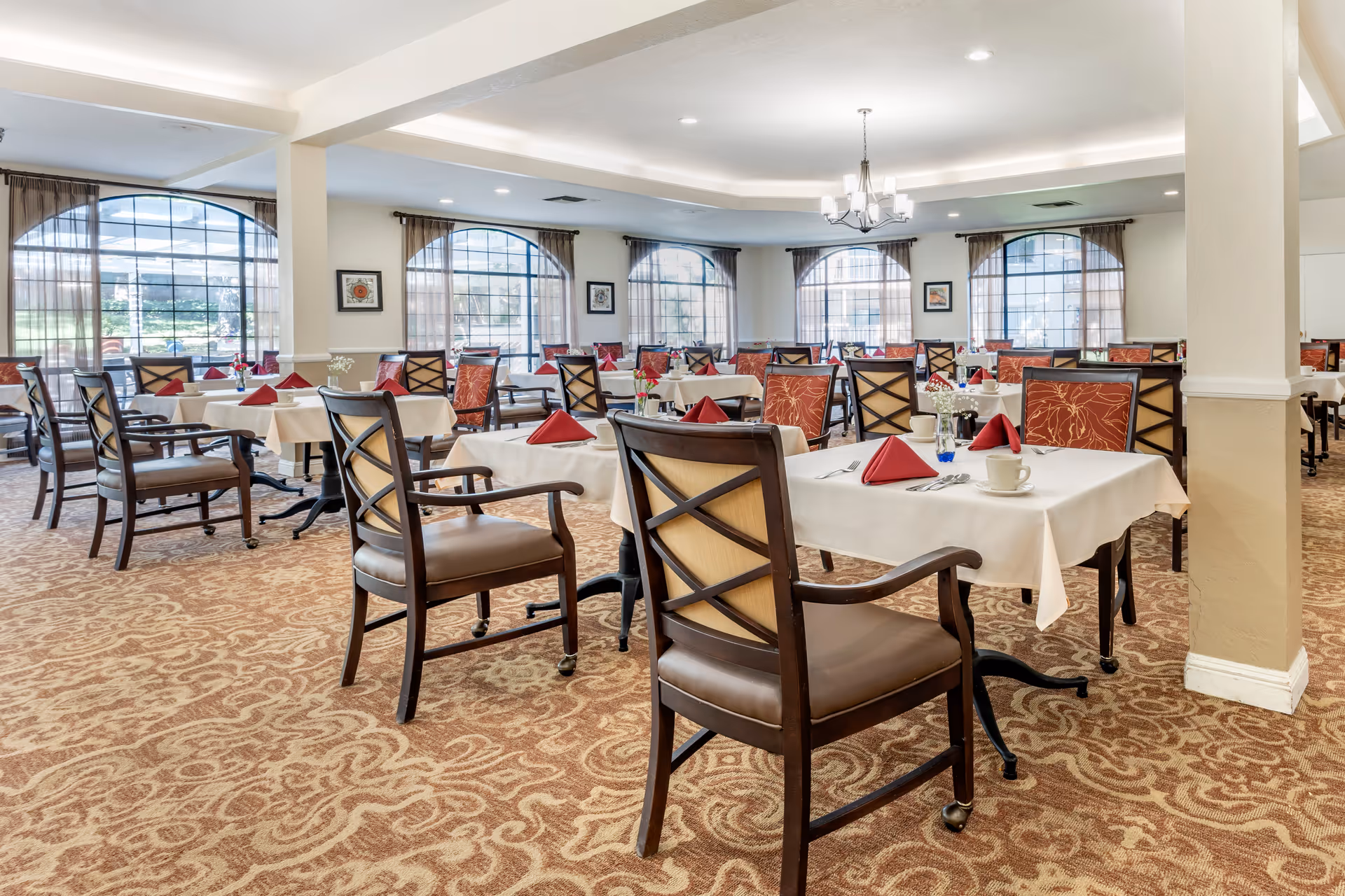 A spacious dining room with multiple tables covered in white tablecloths, each set with red folded napkins, cups, and silverware. The room features large arched windows with sheer curtains, patterned carpet, and wooden chairs with cushioned seats and backs. A chandelier hangs from the ceiling, providing soft lighting.