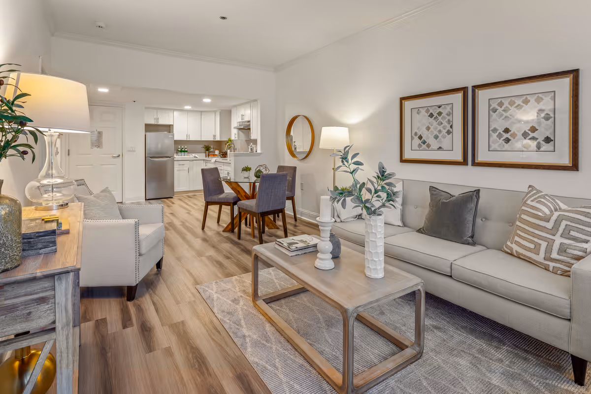 A modern and cozy living room and kitchen area in a senior living facility. The living room features a light gray sofa with decorative pillows, a wooden coffee table with vases and books, a floor lamp, and framed artwork on the wall. Adjacent to the living room is a small dining area with a round wooden table and four upholstered chairs. The kitchen in the background has white cabinetry, stainless steel appliances including a refrigerator, and a clean countertop with some decorative items. The flooring is wood-style laminate, and the overall color scheme is neutral and inviting.