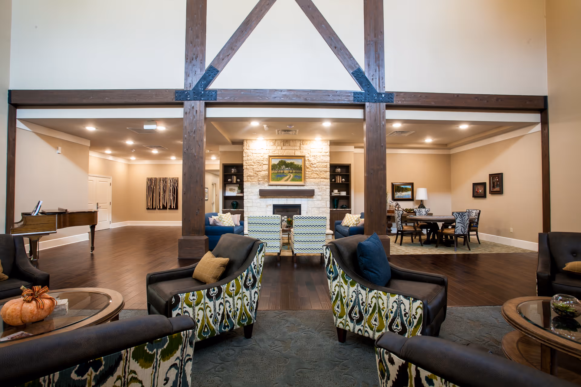 A spacious and well-lit living room area in a senior living facility featuring comfortable armchairs with patterned upholstery, a stone fireplace with a painting above it, a grand piano on the left, and a dining table with chairs on the right. The room has wooden beams, hardwood floors, and soft beige walls with framed artwork.
