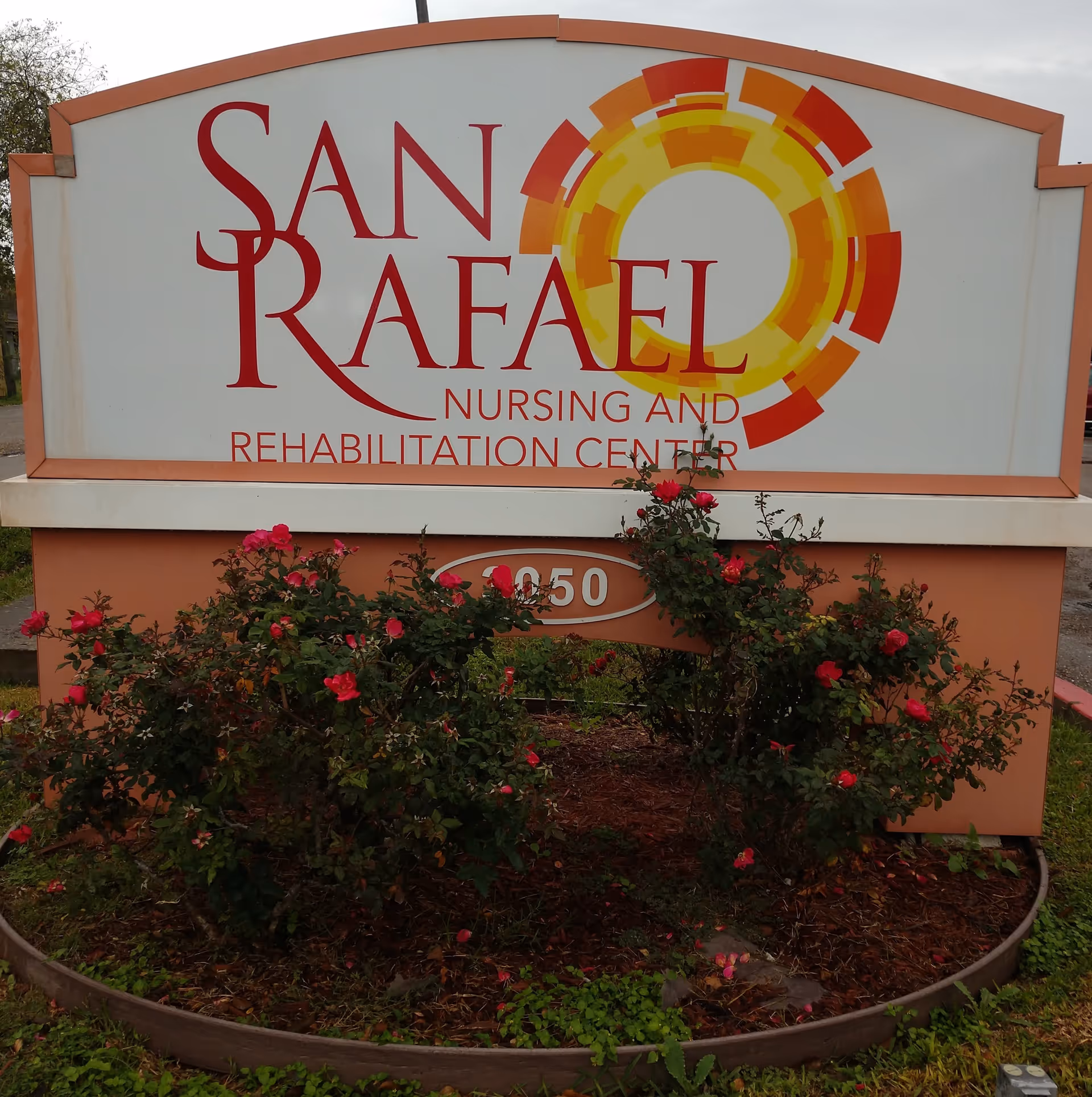 Outdoor sign for San Rafael Nursing and Rehabilitation Center with a decorative circular design in red, orange, and yellow. The sign is surrounded by a flower bed with blooming red roses.