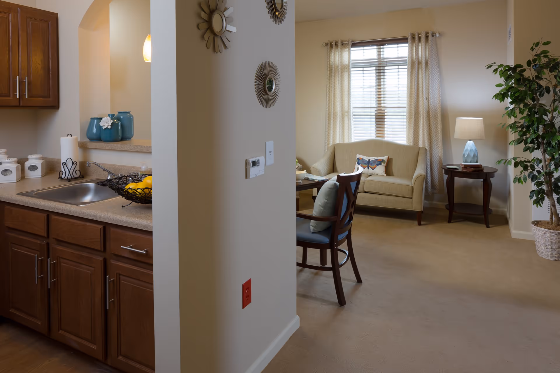 Interior view of a senior living facility showing a small kitchen area with wooden cabinets, a sink, and countertop with decorative jars and a bowl of lemons. Adjacent to the kitchen is a living room with a beige loveseat, a wooden chair with a cushion, a side table with a lamp, a large window with curtains, and a potted plant in the corner.