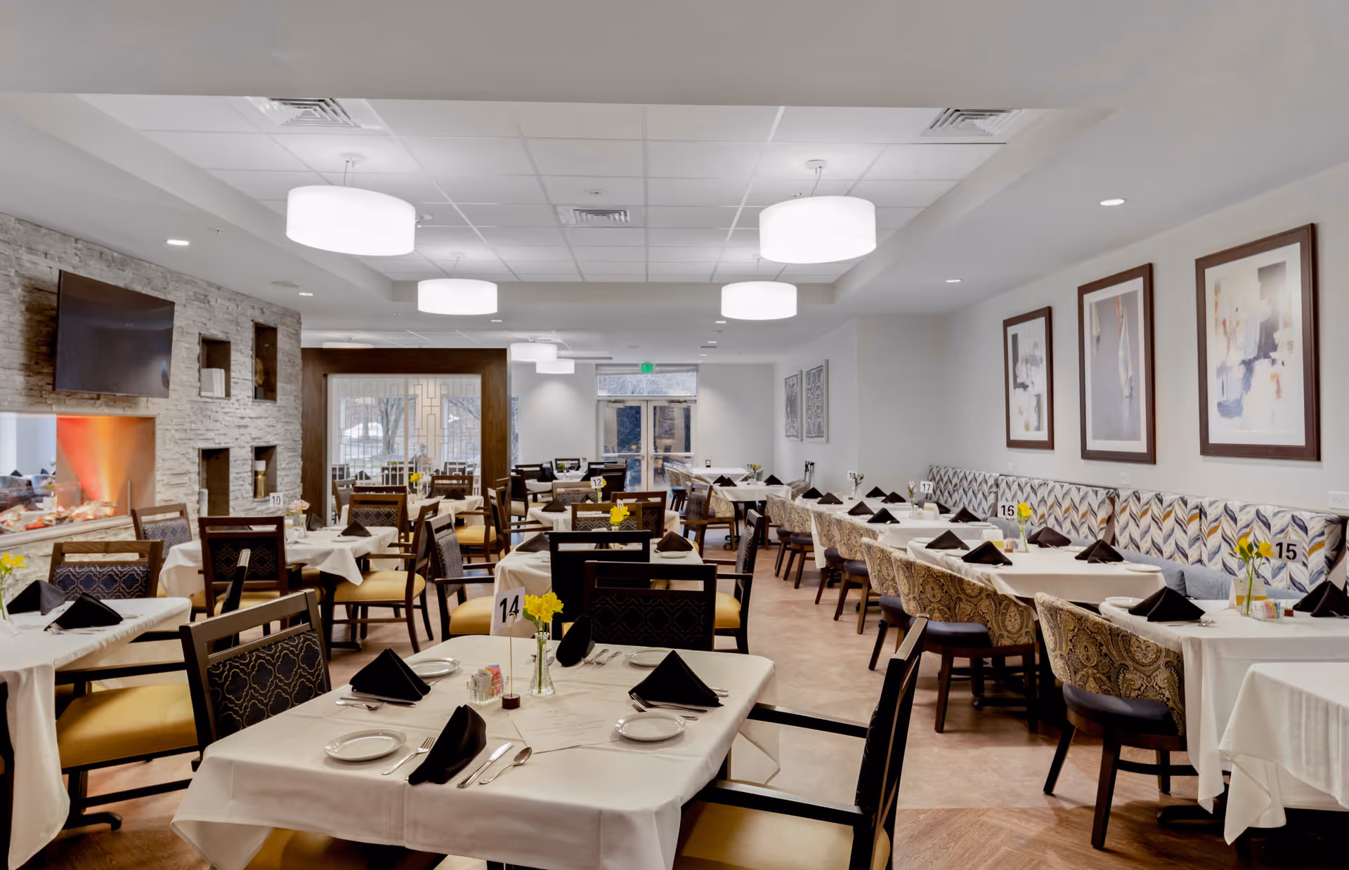 A well-lit dining room with multiple tables covered in white tablecloths, each set with black napkins, plates, and silverware. The room features modern light fixtures hanging from the ceiling, framed abstract artwork on the walls, and a stone accent wall with a mounted TV. Chairs with patterned upholstery and yellow cushions surround the tables, and small vases with yellow flowers decorate each table.