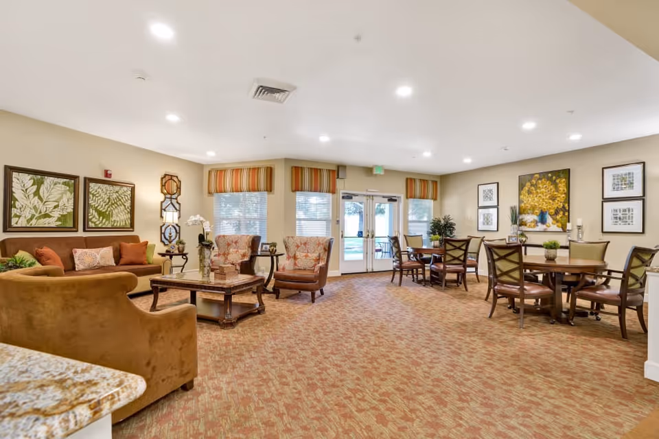 Spacious senior living common room with sofas, armchairs, a coffee table, and dining tables near glass doors.
