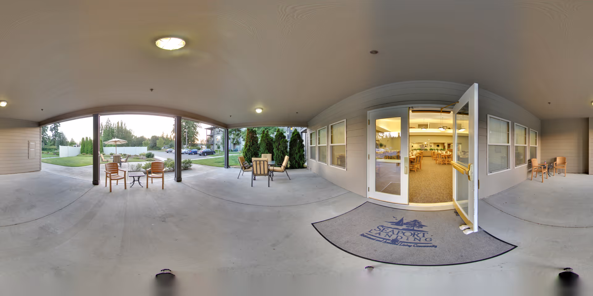 Covered outdoor patio area with several chairs and small tables, adjacent to a building entrance with glass double doors leading into a dining room with wooden tables and chairs. The patio overlooks a grassy area with trees and a parking lot in the background.