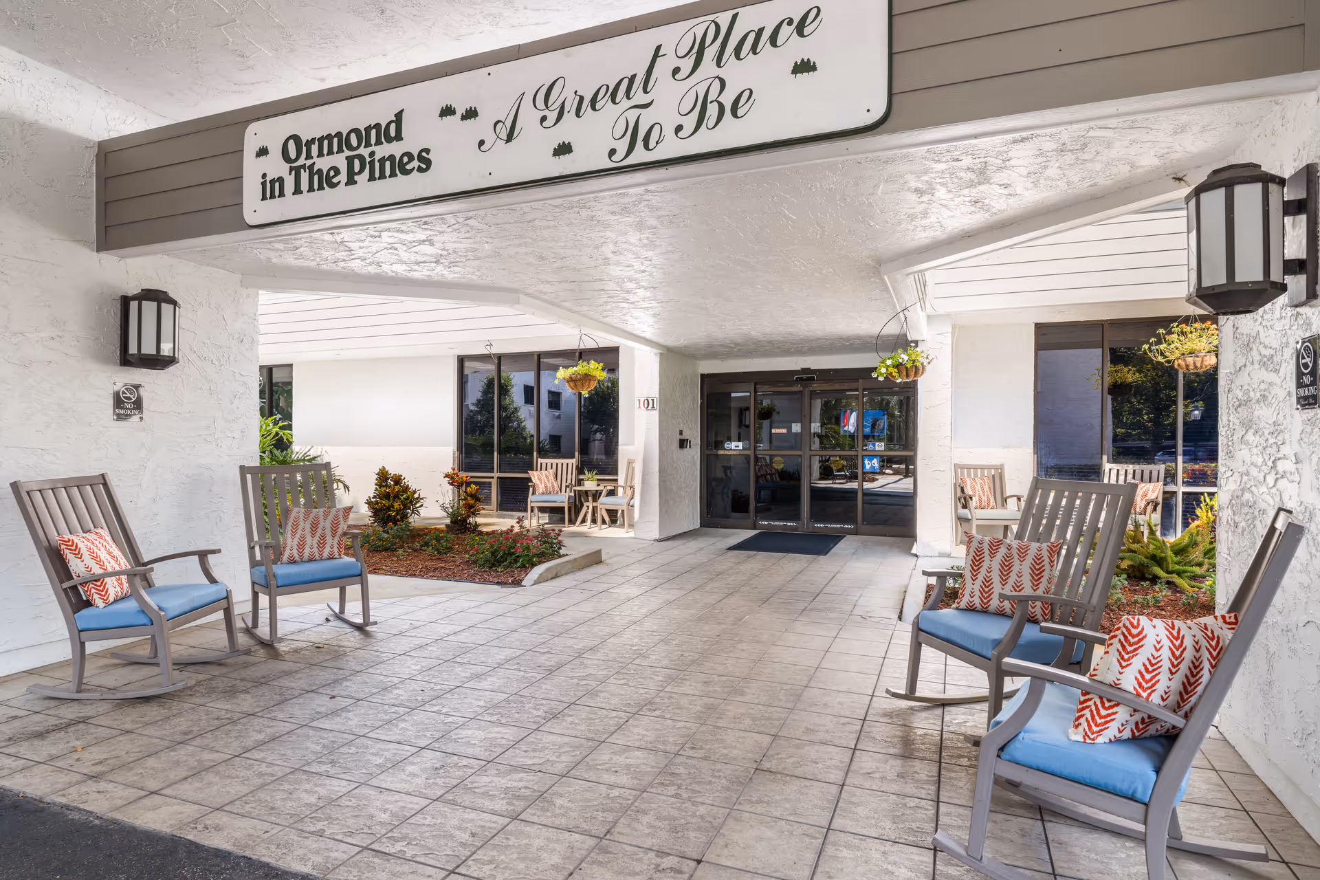 Covered front entry of a senior living facility with rocking chairs, cushions, hanging plants, and a sign reading "A Great Place To Be" above glass doors.