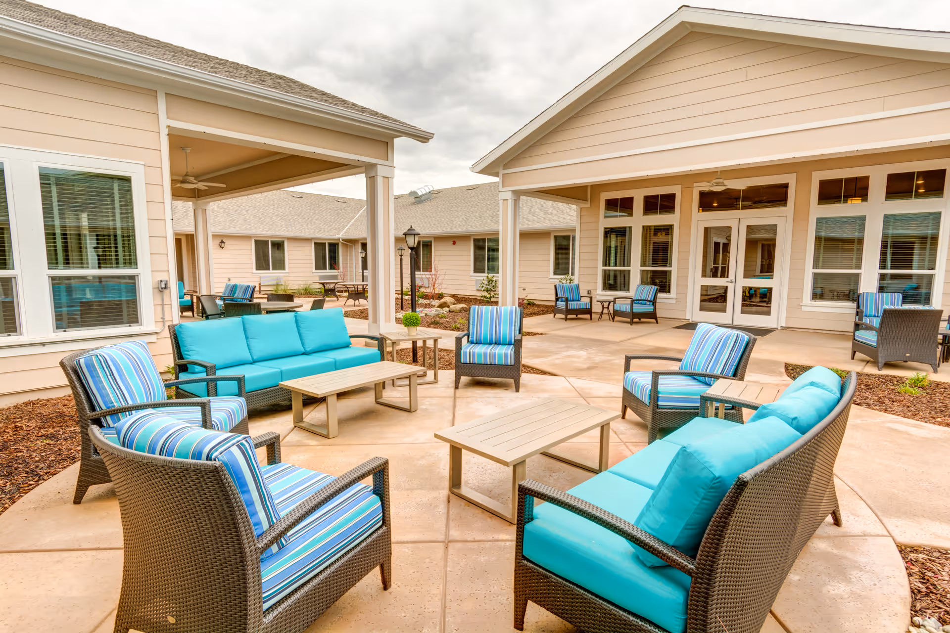 Outdoor seating area at Carmel Village Memory Care featuring wicker furniture with turquoise cushions and striped pillows arranged around wooden coffee tables on a concrete patio, surrounded by beige buildings with large windows and doors under a cloudy sky.