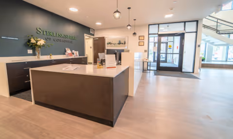 Reception area of Stirlingshire of Coralville featuring a modern front desk with a white countertop and dark wood base. Behind the desk is a dark green wall with the facility name in green letters. The area is well-lit with pendant lights hanging from the ceiling. To the right, there is a glass entrance door with windows allowing natural light to enter the space. The floor is light wood, and there are some decorative items and framed pictures on the walls.