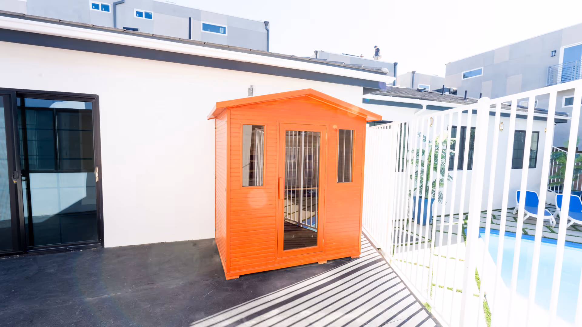 A small orange wooden shed with glass windows and a door, placed on a black patio floor next to a white building wall and a white metal fence. In the background, there are modern buildings and a swimming pool area with blue lounge chairs and potted plants.