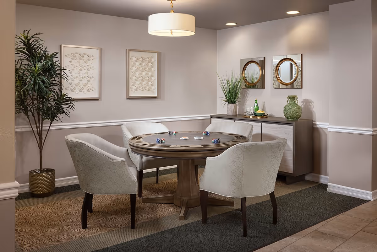 A cozy interior room with a round wooden table surrounded by four upholstered chairs. The table has poker chips and playing cards on it. The room features a sideboard with decorative items including a green vase, a plant, and two round mirrors on the wall above it. There are two framed artworks on the adjacent wall and a tall potted plant in the corner. The lighting is warm with a ceiling light fixture above the table.