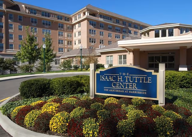Exterior view of the Isaac H. Tuttle Center at The Amsterdam at Harborside, showing a multi-story building with windows, a driveway, landscaped bushes, and a flower bed with yellow and red flowers in front of a blue sign.