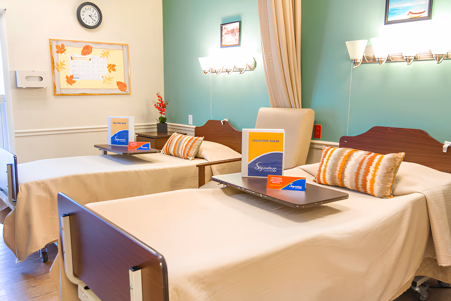 A nursing center room with two hospital-style beds, each with a beige bedspread and a striped pillow. Each bed has an overbed table with informational brochures. The walls are painted light green and white, with framed artwork and a clock on the wall. There is a beige chair between the beds and wall-mounted lights above each bed.