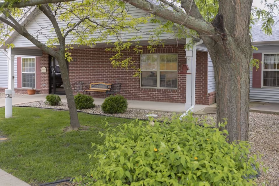 Front porch area of a residential building with a brick wall, two windows, a bench with two chairs, and a tree with green leaves in the foreground. There is a small garden with bushes and a birdhouse attached to a white downspout.