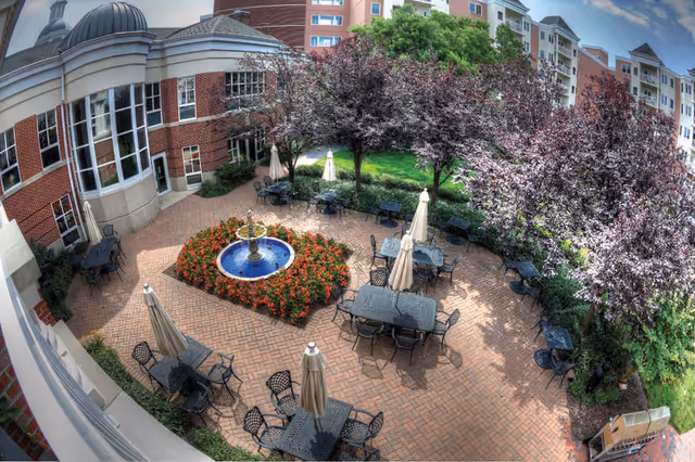 Outdoor brick courtyard with a central fountain surrounded by flower beds, patio tables and umbrellas, trees, and the adjacent multi-story building.