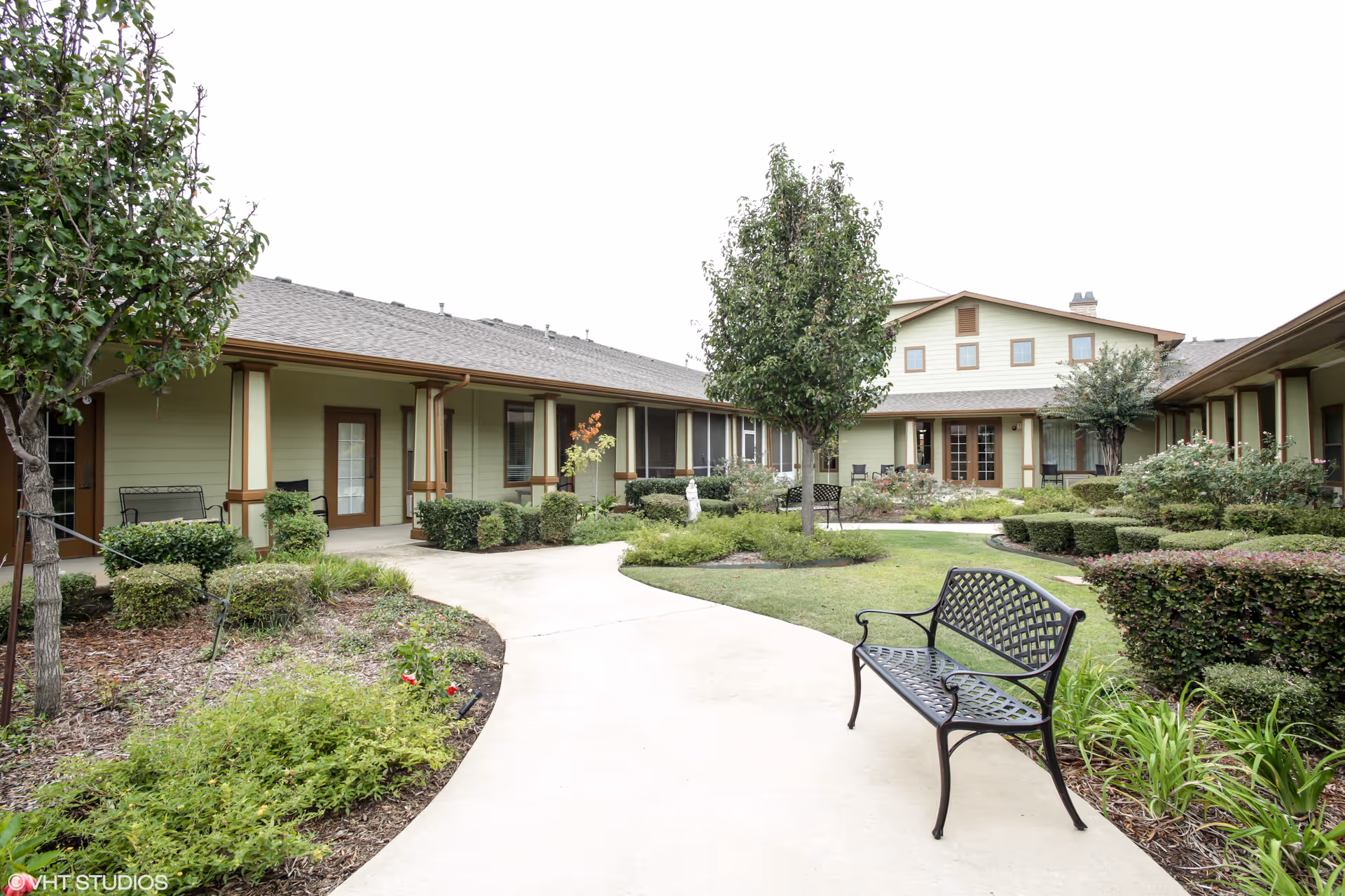 Outdoor courtyard area of Spring Lake Assisted Living and Memory Care featuring a paved walkway, green shrubs, trees, and a black metal bench. The building surrounds the courtyard with light green walls and brown trim.