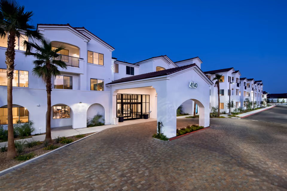 Dusk exterior of Revel Vegas senior living campus with white stucco buildings, arches, and palm trees.