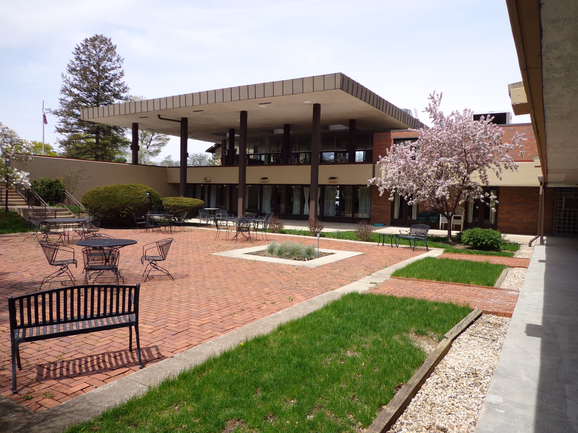 Outdoor courtyard area of Plum Landing Retirement Community featuring brick-paved patio with black metal tables, chairs, and benches. There are green grass patches, a blooming tree with pink flowers, and a modern building with large windows and a flat roof supported by columns in the background.