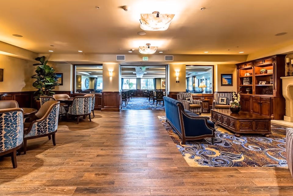 A spacious and well-lit senior living common area featuring wooden flooring and a patterned carpet. The room includes a seating area with a blue sofa, patterned armchairs, a wooden coffee table, and a large wooden bookshelf. There are also several small round tables with upholstered chairs along the left side. The walls are decorated with framed artwork and the ceiling has modern light fixtures. Large windows in the background allow natural light to enter the room.