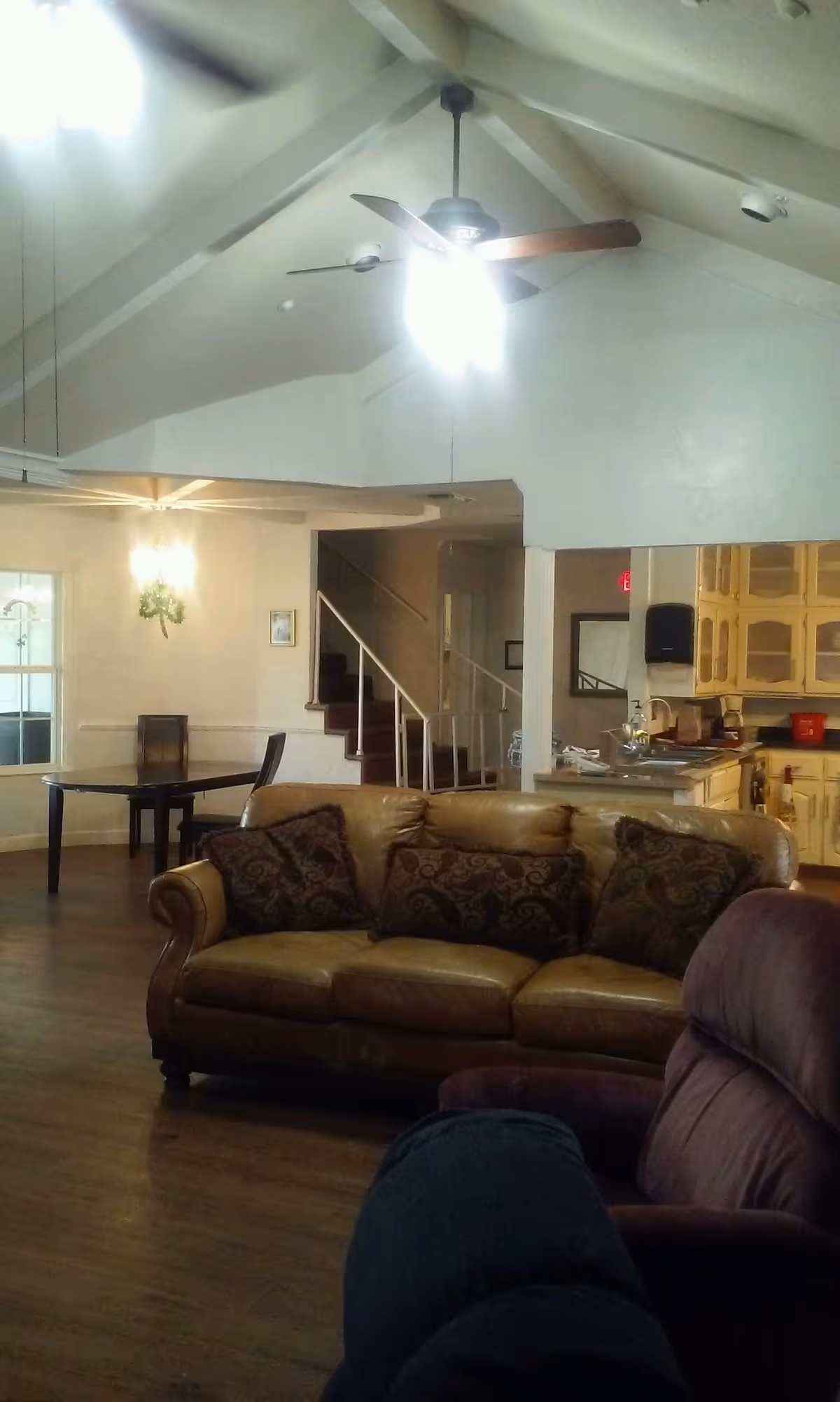 Interior view of a living area in an assisted living facility featuring a brown leather sofa with patterned cushions, a purple recliner chair, a wooden dining table with chairs, a ceiling fan with light, and a kitchen area with cream-colored cabinets in the background. There is a staircase leading to an upper floor and wooden flooring throughout.