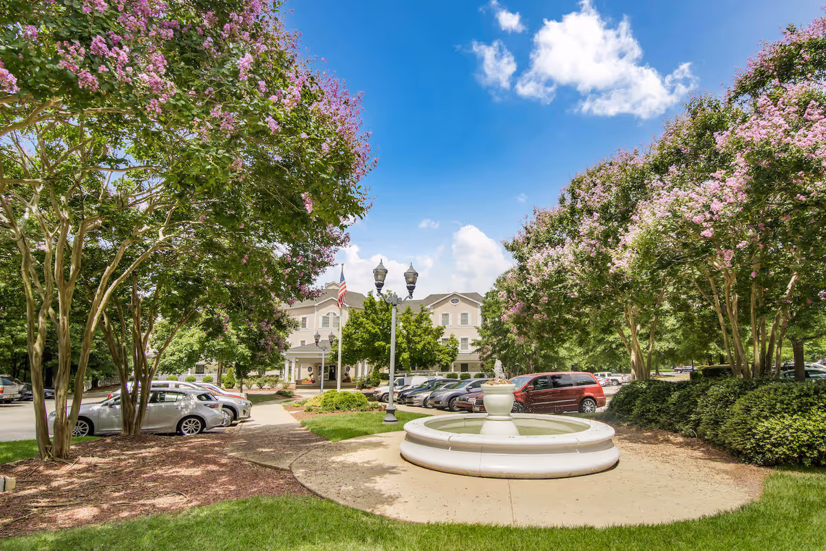 Outdoor view of HeartFields at Cary facility showing a circular water fountain in the foreground, surrounded by green grass and flowering trees with pink blossoms. In the background, there is a parking lot with several cars and a multi-story building under a blue sky with some clouds.