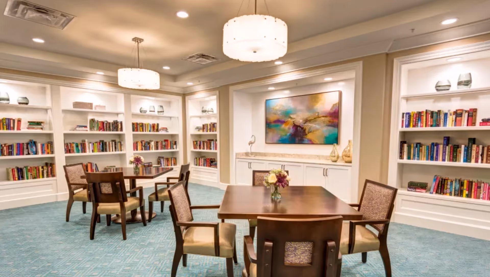 Well-lit library-style common room with built-in bookshelves, wooden tables and chairs, colorful artwork, and hanging light fixtures.