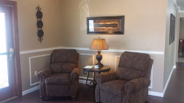 A cozy sitting area with two patterned brown armchairs separated by a small round wooden table holding a lamp and some items. The wall behind is beige with white trim and decorated with a framed artwork and a vertical metal wall ornament. To the left is a wooden door with glass panels, and a hallway extends to the right.