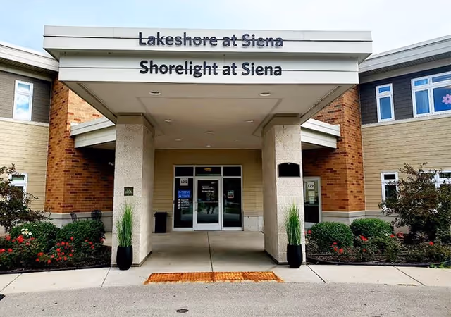 Front entrance of a senior living facility with a covered drop-off area supported by two columns. The building exterior features brick and beige siding with windows. There are two black planters with tall green plants on either side of the entrance. Above the entrance, signs read 'Lakeshore at Siena' and 'Shorelight at Siena'.