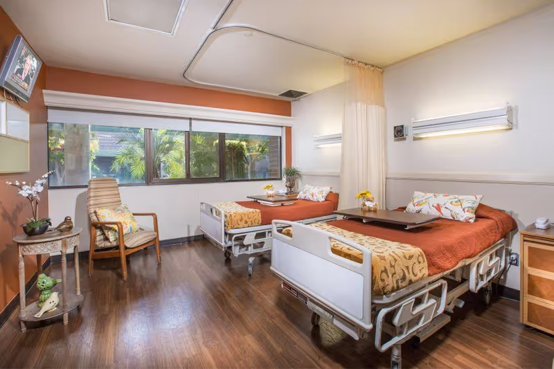 A bright and clean healthcare facility room with two hospital beds, each made with orange bedspreads and decorative pillows. There is a wooden chair with a cushion, a small round table with decorative items, and large windows letting in natural light. The room has wood flooring and a privacy curtain between the beds.
