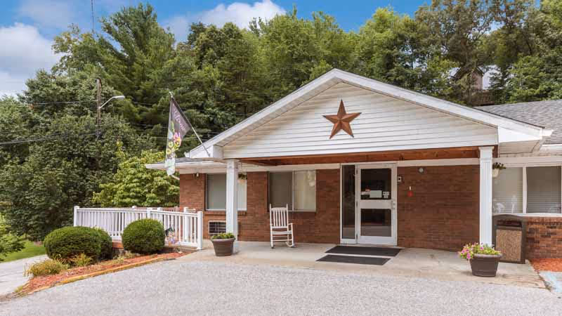 Front entrance of a single-story brick building with a covered porch, rocking chair, potted plants, a decorative star above the door, and trees in the background.