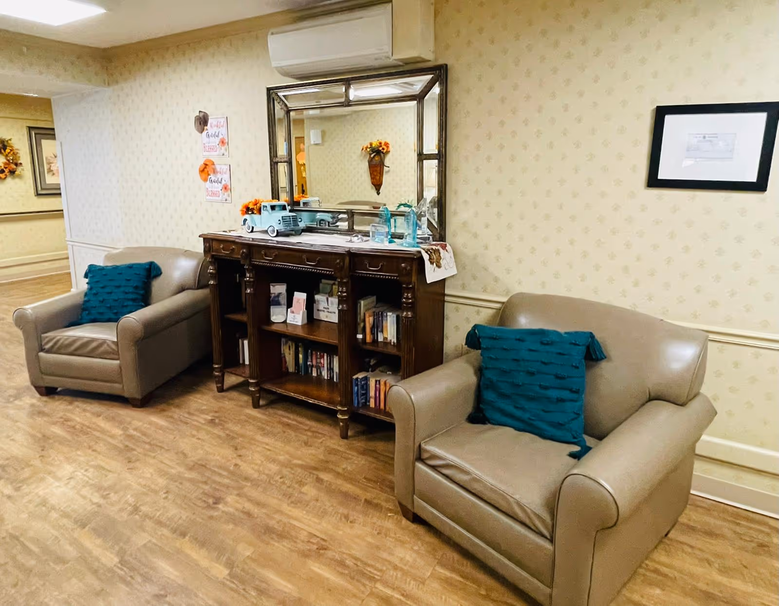 A cozy seating area in a hallway with two beige armchairs, each with a teal cushion. Between the chairs is a wooden console table with shelves holding books and decorative items, topped with a large mirror and small decorative pieces. The walls have light patterned wallpaper and framed artwork, with wood flooring beneath.