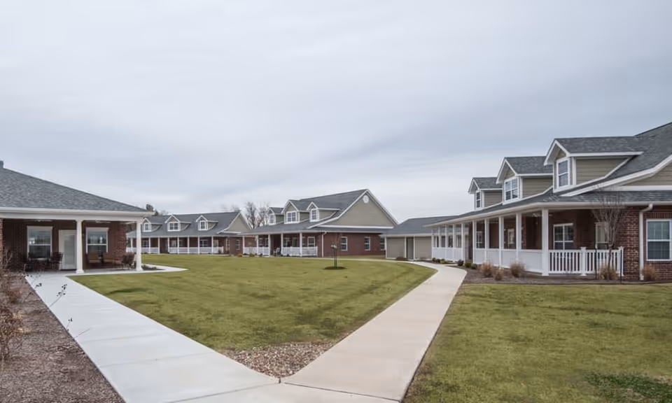 Outdoor view of a senior living facility with multiple single-story and two-story buildings featuring brick and beige siding, white railings, and covered porches surrounding a large grassy courtyard with paved walkways.