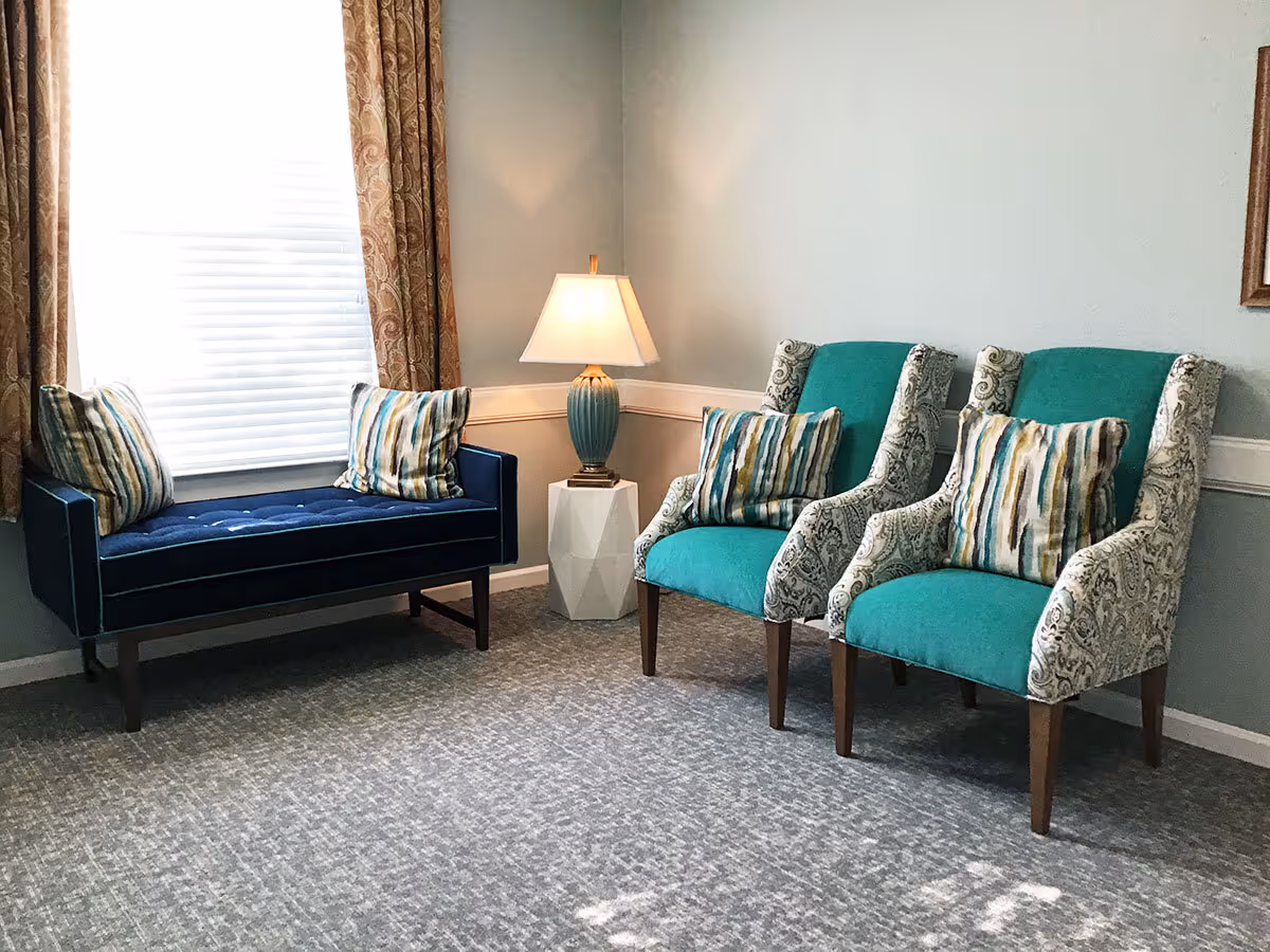 A cozy sitting area with a blue cushioned bench with two striped pillows on the left, two teal armchairs with patterned sides and striped pillows on the right, and a small white side table with a blue lamp in the corner. The room has light gray walls, a window with patterned curtains, and a gray carpeted floor.