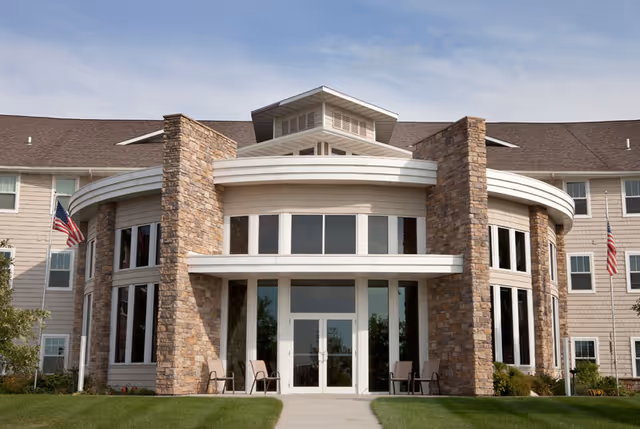 Front exterior view of a senior living facility building with a curved stone facade, large windows, and a central entrance with glass doors. Two American flags are displayed on flagpoles on either side of the entrance, and there are chairs placed near the entrance on a well-maintained lawn.