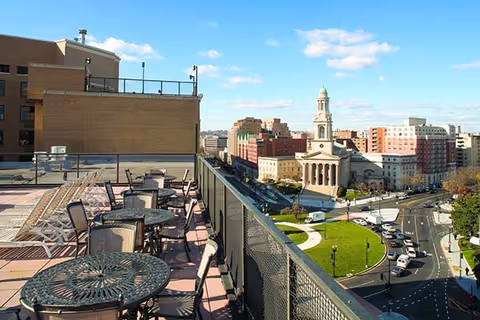 Rooftop patio area with metal tables and chairs overlooking a city street with a church and multiple buildings under a blue sky with scattered clouds.